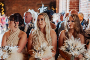 Three women in matching beige dresses sit side by side, each holding a bouquet of white flowers at Hazel Gap Barn. Surrounded by guests, they attend a beautiful Hazel Gap Barn wedding by Cripps and Co.