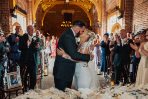 A bride and groom kiss at their wedding ceremony inside the warmly lit Hazel Gap Barn, surrounded by applauding guests. The bride wears a white lace dress, the groom a dark suit, and a loved one’s photo sits on a chair in the front row.