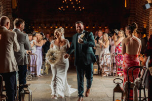 A bride and groom walk down the aisle smiling and holding hands, surrounded by applauding guests in a warmly lit Hazel Gap Barn wedding venue with exposed brick walls and candles—a signature Cripps and Co celebration.