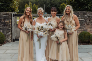 A bride in a white dress stands outdoors with four bridesmaids and a flower girl, all in light beige dresses holding dried flower bouquets, smiling at the camera at a beautiful Hazel Gap Barn wedding. Lush greenery and a stone wall complete the Cripps & Co setting.