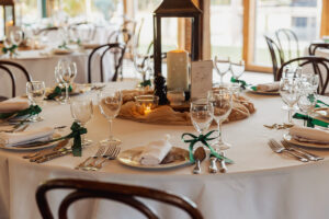 A round table set for a Hazel Gap Barn wedding with white tablecloth, wine glasses, cutlery, napkins tied with green ribbon, a lantern centerpiece, candles, and a table number card. Wooden chairs surround this elegant Cripps & Co. setting.