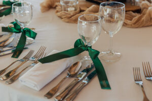 Elegant table setting at a Hazel Gap Barn wedding, featuring white napkins, silver cutlery, and two wine glasses per place. Green ribbons accent each napkin and cutlery, adding charm to the neatly arranged plates.