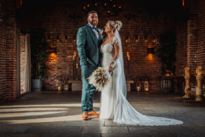 A bride and groom stand together in the warmly lit Hazel Gap Barn, with brick walls and candles. The bride wears a white gown and holds a bouquet, while the groom wears a green suit and brown shoes at this Cripps & Co venue.