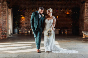 A bride in a white lace gown holding a bouquet walks arm in arm with her groom in a green suit. They smile at each other in the warmly lit Hazel Gap Barn, a rustic Cripps and Co venue adorned with twinkling lights.