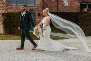 A bride in a long white gown with a flowing veil and a groom in a dark suit walk hand in hand outdoors, smiling, in front of the rustic brick walls of Hazel Gap Barn. The bride holds a bouquet of flowers at their Hazel Gap Barn wedding.