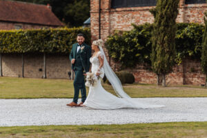A bride in a white gown with a long veil walks outdoors on gravel next to a groom in a green suit. Both are smiling, surrounded by greenery and the charm of historic brick buildings at their Hazel Gap Barn wedding.