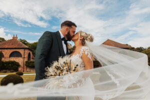 A bride and groom share a kiss outdoors at Hazel Gap Barn, her veil flowing in the wind. She holds a bouquet of dried flowers as they stand before brick buildings and a partly cloudy sky—perfect for a Hazel Gap Barn wedding by Cripps and Co.