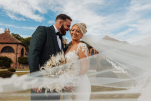 A smiling bride in a white lace gown holds a bouquet, standing beside her groom at Hazel Gap Barn. Her veil flows in the wind, with brick buildings and blue sky capturing the charm of their special day at Hazel Gap.