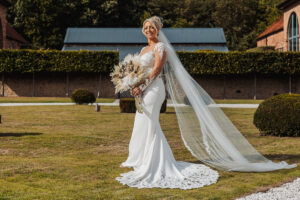 A bride in a white lace wedding dress and long veil stands on a lawn at Hazel Gap Barn, holding a bouquet of dried flowers and smiling, with buildings and greenery from her Hazel Gap Barn wedding in the background.