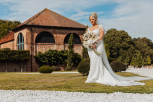 A bride in a white wedding dress holds a bouquet of flowers while walking on grass in front of the rustic brick Hazel Gap Barn by Cripps and Co, basking in the sunshine.