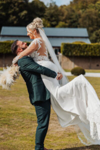 A groom in a dark suit lifts his smiling bride, who wears a long white gown and veil, outdoors on a sunny day at Hazel Gap Barn. The happy couple is surrounded by grass and trees, with the beautiful Cripps & Co venue in the background.