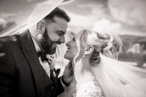 A bride and groom smile lovingly at each other under a flowing veil, their faces close together, outdoors on their hazel gap barn wedding day. The image is in black and white.