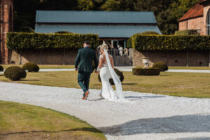A bride in a white dress and veil walks hand in hand with a groom in a dark suit along a gravel path at Hazel Gap Barn, surrounded by landscaped gardens, trimmed bushes, and historic brick buildings from Cripps and Co.