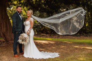 A bride in a white lace gown and long veil stands beside her groom at their Hazel Gap Barn wedding. Her veil flows in the breeze as they smile, standing on grass with trees and the charm of Hazel Gap in the background.