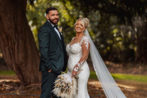 A bride in a white lace wedding dress and a groom in a dark suit stand outdoors at Hazel Gap Barn, smiling and posing together. The bride holds a bouquet of dried flowers, with lush trees and greenery from Cripps & Co as their backdrop.