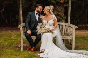 A bride and groom sit closely on a wooden bench outdoors at their Hazel Gap Barn wedding, smiling at each other. The bride holds a dried flower bouquet and wears a white lace dress with a long veil; the groom wears a dark suit with brown shoes.