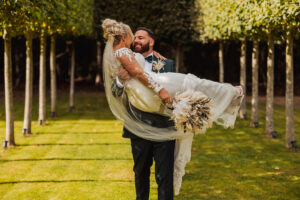 A groom in a dark suit joyfully lifts his smiling bride, who is wearing a white dress and veil, at their Hazel Gap Barn wedding, surrounded by sunlit gardens and rows of trimmed trees. She holds a dried floral bouquet.