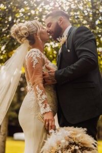 A bride and groom share an intimate moment, leaning in for a kiss outdoors at their Hazel Gap Barn wedding. The bride, in a lace dress with bouquet, and the groom in a dark suit, are framed by sunlight filtering through the trees.