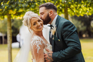 A groom in a dark green suit kisses his smiling bride, who wears a white lace dress and veil, on the temple. They stand outside Hazel Gap Barn under leafy trees on a sunny day, capturing a perfect Cripps and Co moment.