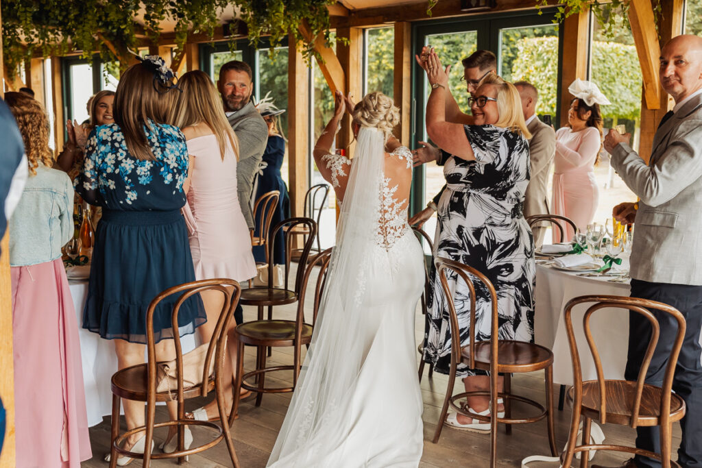 A bride in a white dress walks through a group of standing guests who are clapping and smiling at an indoor reception with wooden chairs and greenery overhead.
