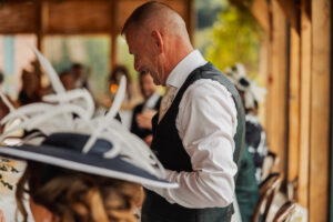 A man in formal attire smiles and looks down while standing indoors at a lively Hazel Gap Barn wedding. Other guests, some wearing decorative hats, mingle in the background as warm light fills the wooden Cripps and Co venue.