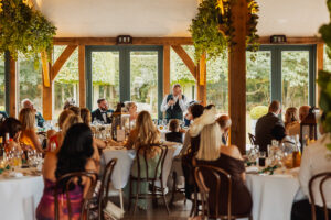 A man in a suit stands, speaking into a microphone, addressing guests seated at round tables in the bright, elegant Hazel Gap Barn wedding venue with wooden beams, greenery, and large windows overlooking gardens.