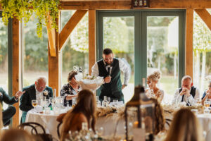 A man in a vest stands and speaks into a microphone at a decorated Hazel Gap Barn wedding reception table, surrounded by seated guests, some smiling and others looking down, with greenery and large windows in the background.