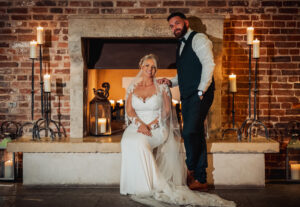 A bride in a white gown sits smiling by the fireplace at her Hazel Gap Barn wedding, while the groom stands beside her with a hand on her shoulder. Warm candlelight and the rustic brick wall create a cozy Cripps and Co atmosphere.