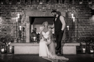 A bride sits on a stone hearth in front of a brick fireplace at Hazel Gap Barn, smiling up at the groom beside her. Both in wedding attire, surrounded by lit candles and lanterns—the timeless scene is captured in black and white.