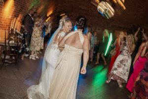 Two women in elegant dresses, one in a white wedding gown and veil, dance joyfully together at a lively Hazel Gap Barn wedding, surrounded by guests in colorful attire and blurred lights at this charming Cripps and Co venue.