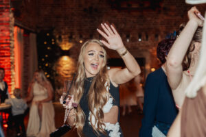 A young woman with long blonde hair waves and smiles at the camera while holding a drink at a lively hazel gap barn wedding, with people and warm lights in the background.