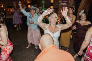 A bride in a white lace wedding dress dances with her hands raised at a lively indoor Hazel Gap Barn wedding, surrounded by smiling guests in colorful outfits. Everyone enjoys the vibrant atmosphere of this unforgettable Cripps and Co celebration.