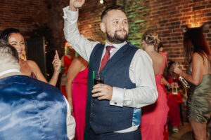 A man in a suit vest with a red tie dances and holds a drink at a lively Hazel Gap Barn wedding, surrounded by people in colorful dresses, with brick walls and warm lighting in the background.