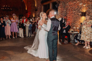 A bride and groom share their first dance at a Hazel Gap Barn wedding, surrounded by smiling guests who watch and take photos. Warm lighting and exposed brick walls create the cozy atmosphere Cripps & Co is known for.