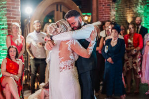 A bride and groom share a tender embrace while dancing at their Hazel Gap Barn wedding, surrounded by guests in formal attire. Warm lights and exposed brick walls create a festive atmosphere.