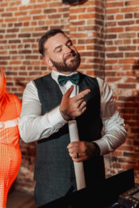 A bearded man in a vest, white shirt, and bow tie smiles and gestures with one hand while holding a rolled-up paper, standing in front of a brick wall at a Hazel Gap Barn wedding by Cripps and Co.