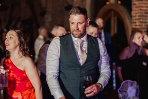A man in a suit vest and tie holds a drink at a social event, surrounded by guests. Beside him, a woman in a red dress also holds a drink. The dimly lit background hints at the rustic charm of a Hazel Gap Barn wedding, with blurred people and brick walls.