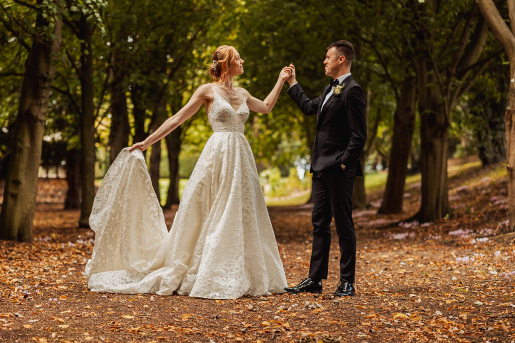 A bride in a white gown and a groom in a black suit hold hands and pose along Saltmarshe Hall’s tree-lined, leafy pathway, surrounded by warm autumn colors.