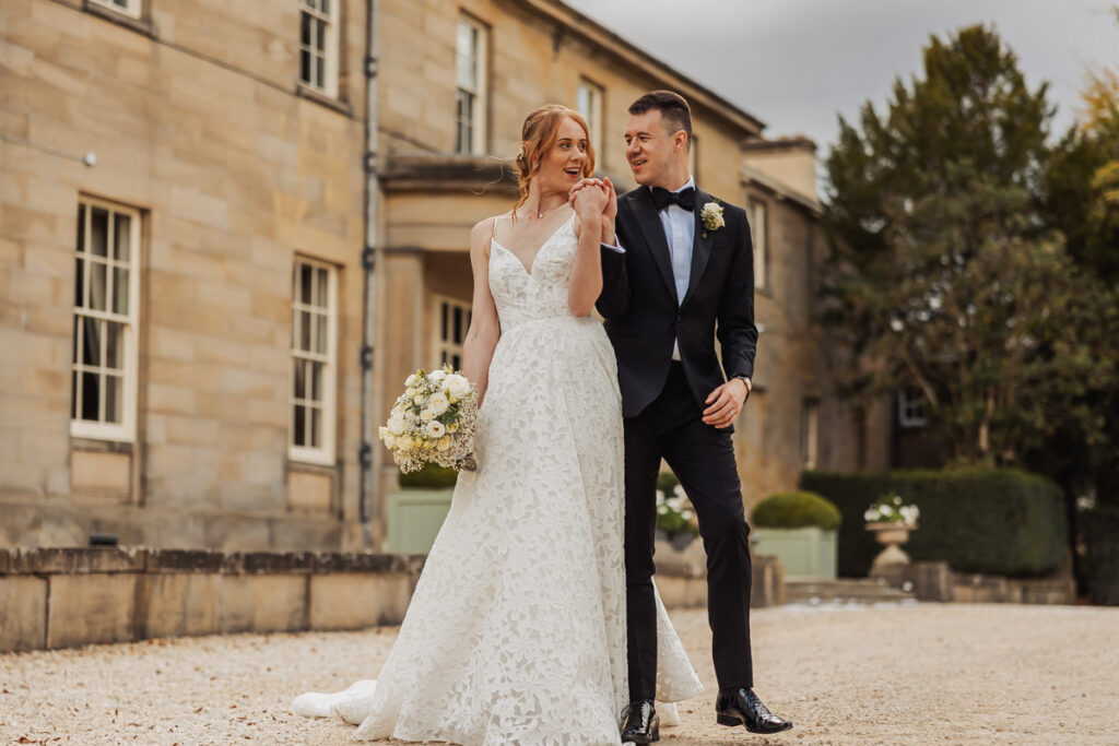 A bride in a white lace gown holds a bouquet and a groom in a black tuxedo walk together, smiling and holding hands, outside Saltmarshe Hall's grand stone building on a cloudy day.