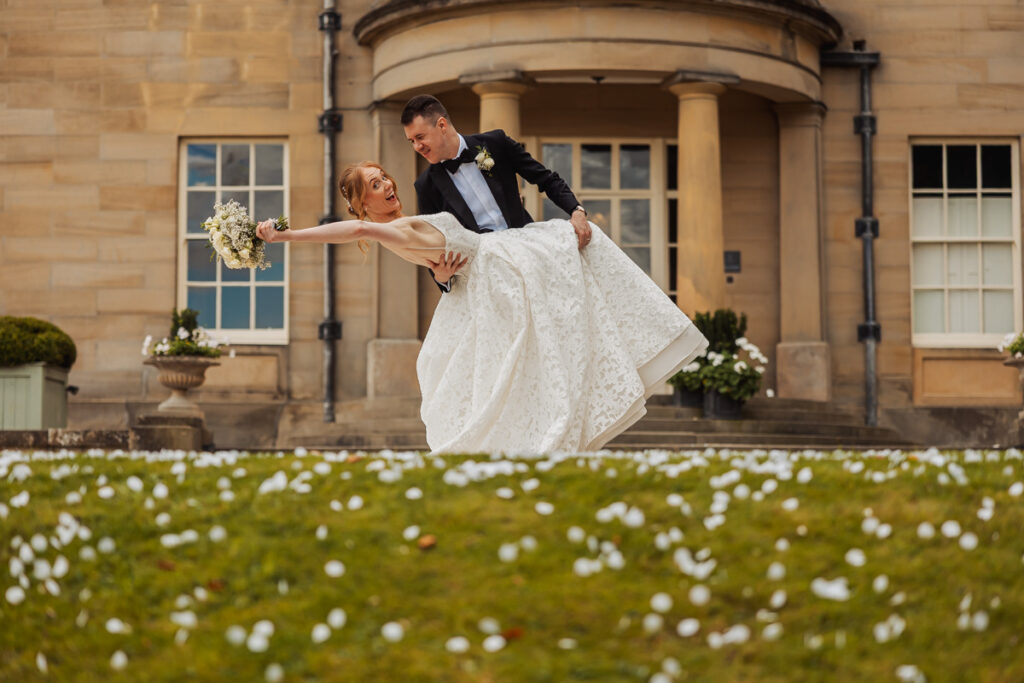A groom in a black suit lifts his smiling bride in a white dress outside a grand building, surrounded by scattered flower petals on the grass. The bride holds a bouquet, and both look joyful 