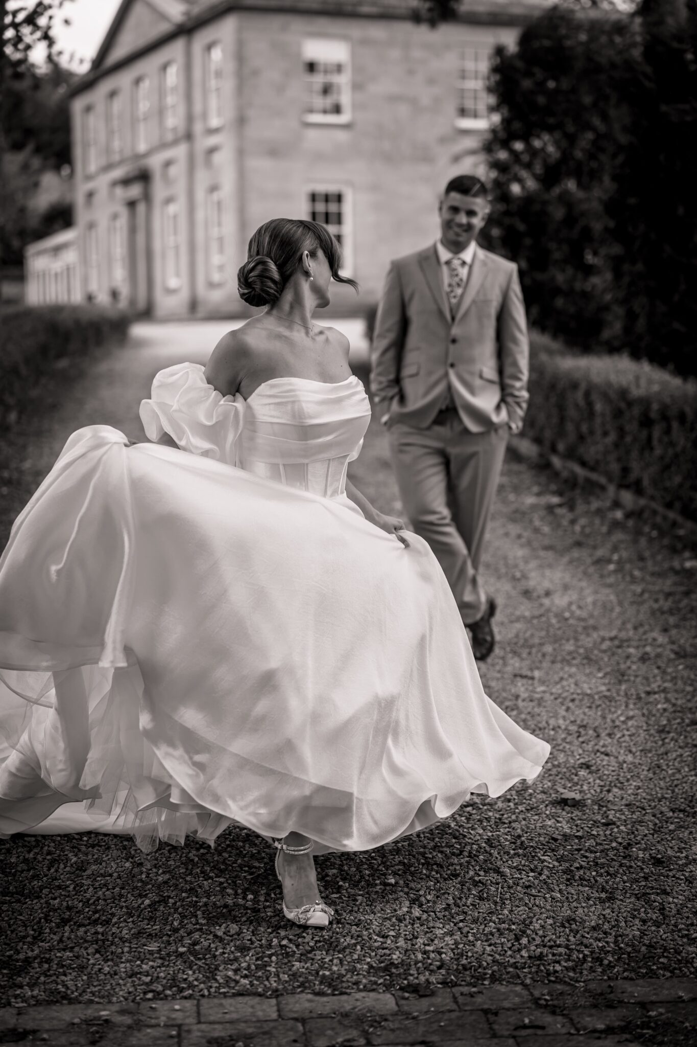 A bride in a flowing gown walks ahead of her groom along a gravel path in front of a grand stately home, a black and white Yorkshire wedding photograph.