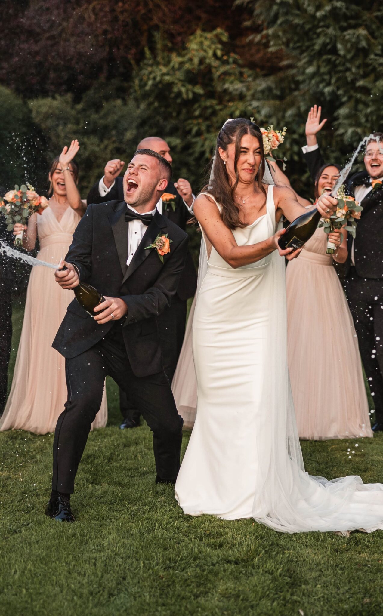 A joyful groom and bride spray champagne together as cheering bridesmaids and groomsmen in blush and black celebrate around them at their fun Yorkshire wedding.