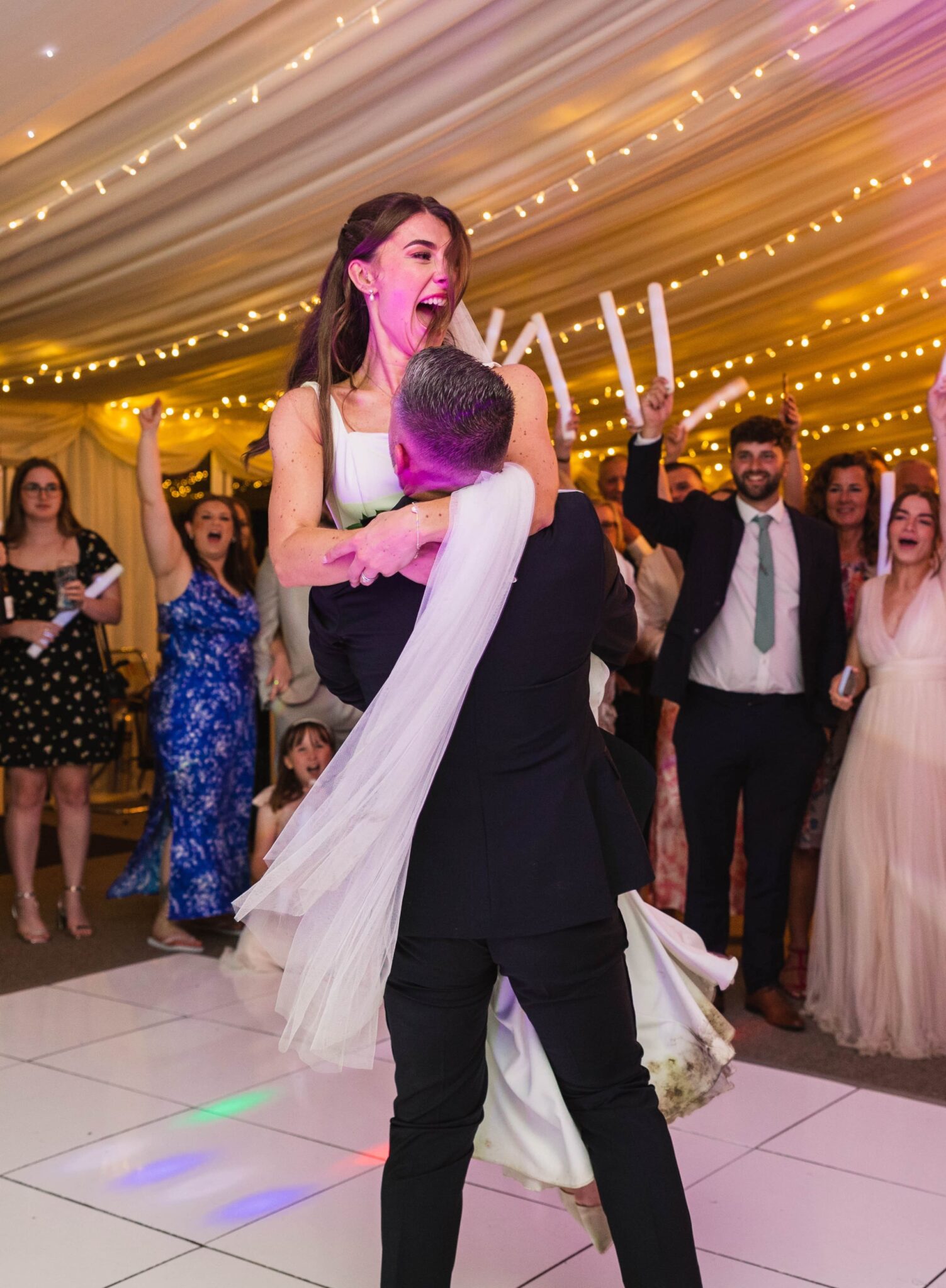 A groom lifts his laughing bride onto the dance floor as wedding guests cheer under fairy lights at their Yorkshire marquee wedding reception.