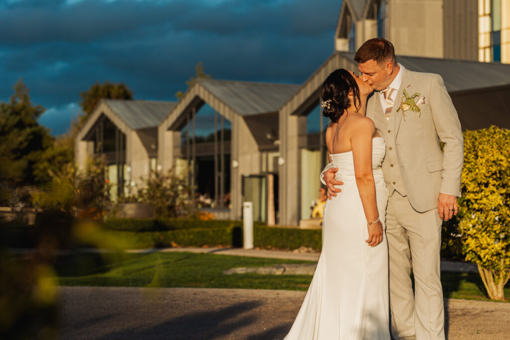 A couple in wedding attire stands close together in front of a modern building with large glass windows, in a warmly lit garden setting