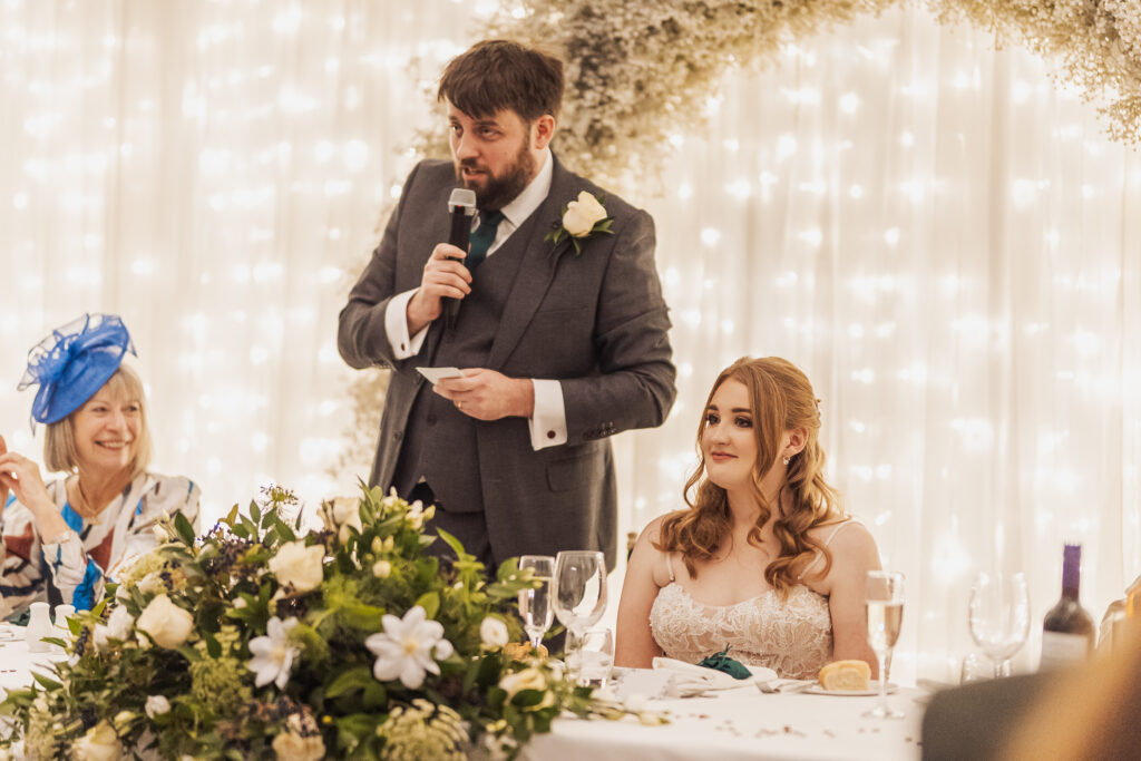 The groom delivers a heartfelt speech while the bride, adorned in a lace gown, sits beside him, surrounded by guests and a floral centerpiece under twinkling fairy lights at Coniston Hotel, Skipton