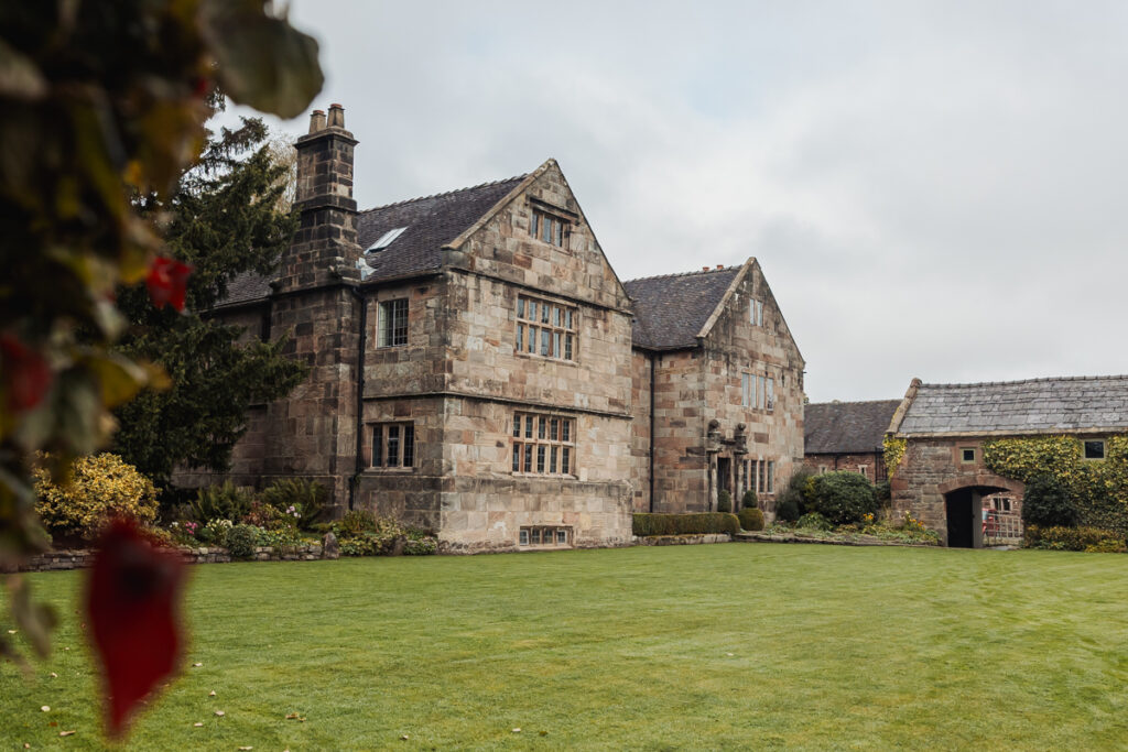 A large historic stone house with gabled roofs and tall chimneys sits next to a neatly manicured lawn in Staffordshire, surrounded by trees and bushes on a cloudy day. Ashes Barn, a charming stone outbuilding, is visible to the right—perfect for a wedding setting.
