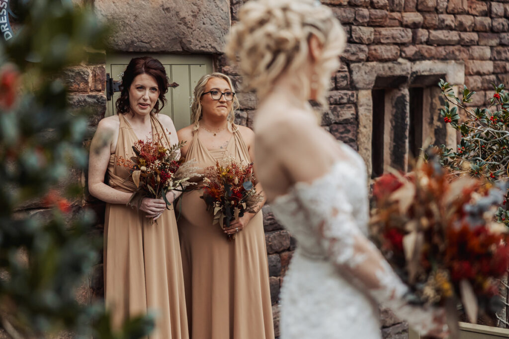 Two bridesmaids in tan dresses holding autumn-themed bouquets stand against a stone wall at Ashes Barn in Staffordshire, looking at a bride in a white lace gown who is slightly out of focus in the foreground.