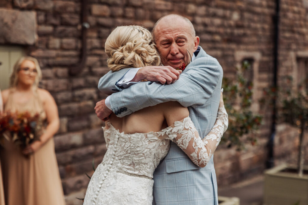 At an Ashes Barn Wedding in Staffordshire, a bride in a white lace dress hugs an older man in a light blue suit outdoors, both with emotional expressions. A bridesmaid holding a bouquet stands blurred in the background near a brick wall.