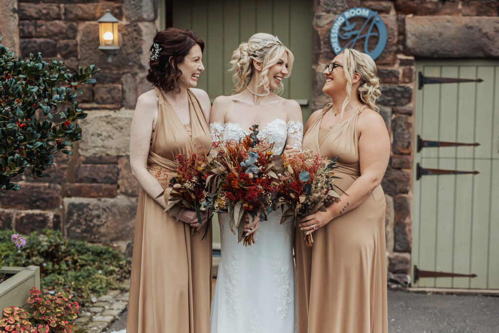 Three women stand smiling outside Ashes Barn in Staffordshire. The bride, in a white dress, is flanked by two bridesmaids in tan gowns. All three hold autumn-themed bouquets and share a joyful wedding moment together.