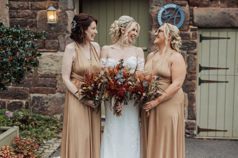 Three women stand smiling outside Ashes Barn in Staffordshire. The bride, in a white dress, is flanked by two bridesmaids in tan gowns. All three hold autumn-themed bouquets and share a joyful wedding moment together.